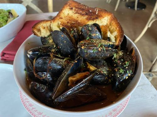 A bowl of mussels in broth, garnished with herbs and spices, served with a slice of toasted bread.