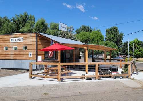 A wooden restaurant with a patio, featuring an umbrella and an American flag, set against a clear blue sky.