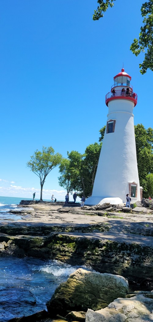 A white lighthouse with a red top stands by the shore, surrounded by trees and people enjoying the sunny day.