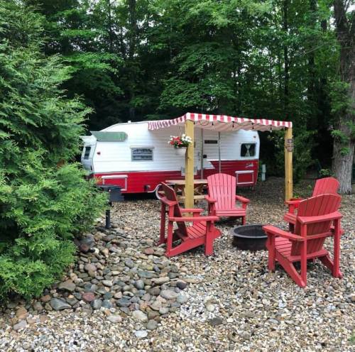 A vintage red and white camper surrounded by trees, with red Adirondack chairs and a fire pit nearby.