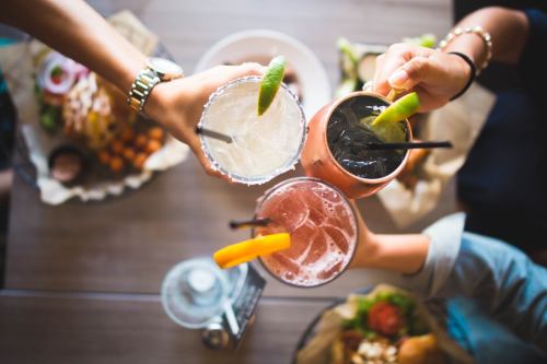 Four hands holding colorful cocktails with garnishes, set on a table with food in the background.