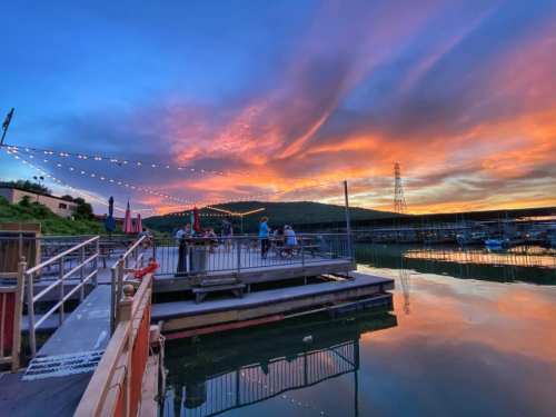 A serene lakeside scene at sunset, with colorful skies reflecting on the water and string lights illuminating a dock.