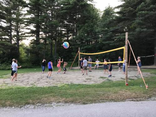 A group of people playing volleyball on a sandy court surrounded by trees during sunset.