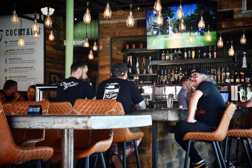 A lively bar scene with patrons sitting at a wooden counter, enjoying drinks and watching a TV screen in a rustic setting.