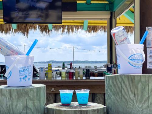 Two buckets with drinks sit on wooden posts, with a bar and scenic view in the background under a colorful awning.