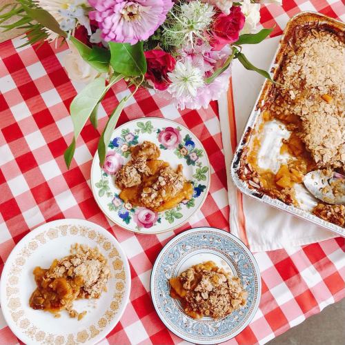 A table with a checkered cloth featuring three plates of apple crisp and a floral centerpiece.