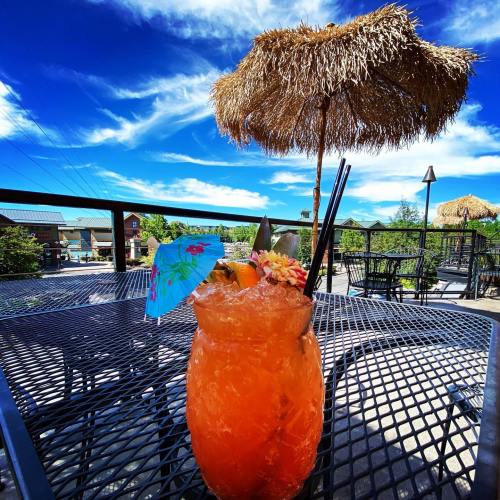 A colorful tropical cocktail with an umbrella and fruit garnish, set on a patio with a blue sky and straw umbrellas in the background.