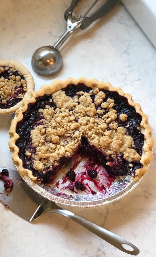 A berry pie with a crumbly topping, partially sliced, on a marble countertop with a pie server and scoop nearby.