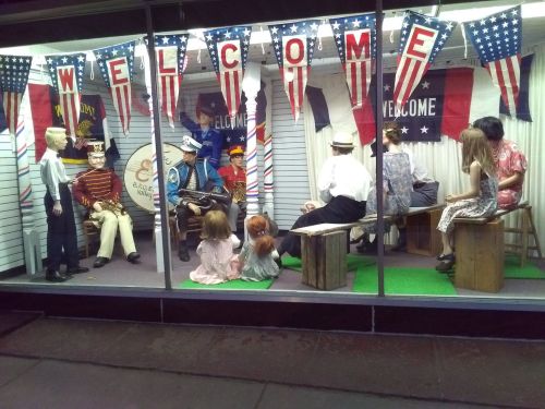 A storefront display featuring mannequins in a patriotic setting, with flags and a band scene.