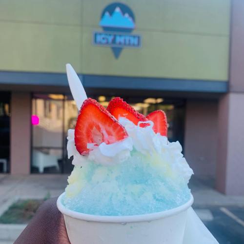A hand holding a cup of colorful shaved ice topped with whipped cream and strawberries, with a shop in the background.