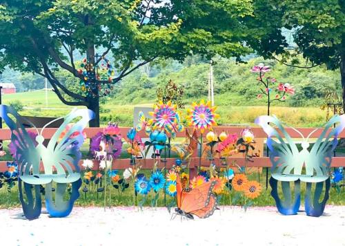Colorful metal flowers and butterfly sculptures in a garden setting, with trees and a scenic background.