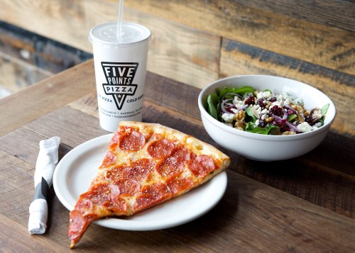 A slice of pepperoni pizza on a plate, alongside a salad and a drink in a cup on a wooden table.