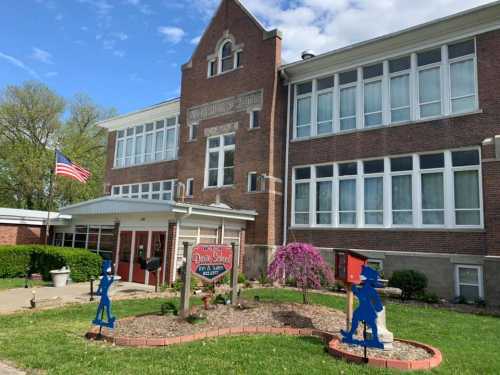 Historic brick building with large windows, American flag, and colorful garden decorations in front.