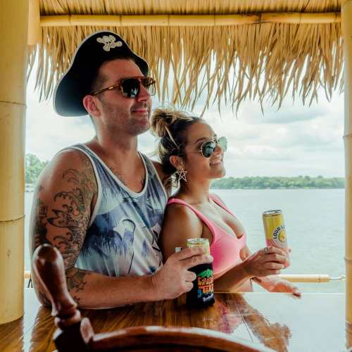 A man in a pirate hat and a woman in sunglasses enjoy drinks by the water, smiling and posing together.