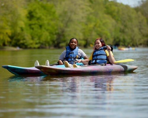 Two young people in colorful kayaks, smiling and enjoying a sunny day on a calm river surrounded by greenery.