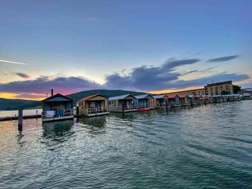 Colorful houseboats lined up on calm water at sunset, with hills and clouds in the background.