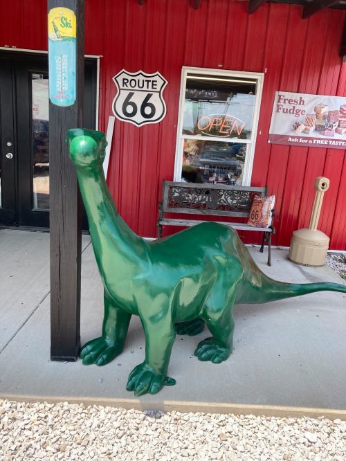 A green dinosaur statue stands outside a building with a Route 66 sign and an "Open" neon sign.