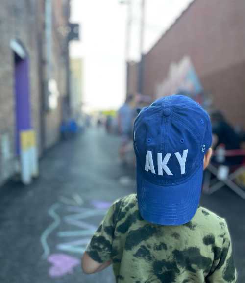 A child wearing a blue cap with "AKY" on it, standing in a colorful alley with people in the background.