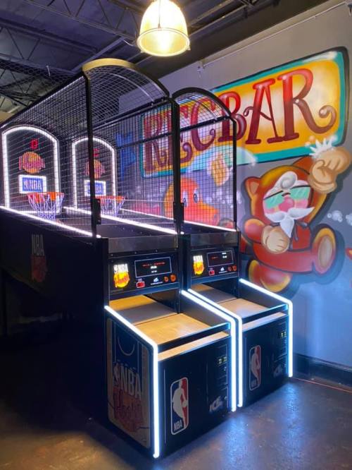 A brightly lit NBA basketball arcade game with a colorful mural in the background featuring a cartoon bear.