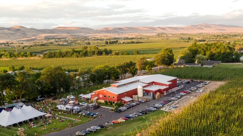 Aerial view of a vineyard with a red barn, cars parked, and people gathered outdoors against a backdrop of rolling hills.