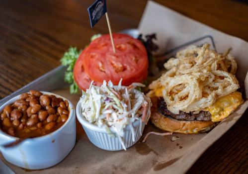 A burger topped with crispy onions, served with coleslaw, baked beans, and fresh tomato on a tray.