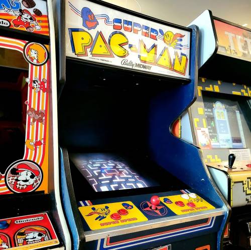 An arcade machine for "Super Pac-Man" featuring colorful graphics and a joystick, surrounded by other arcade games.