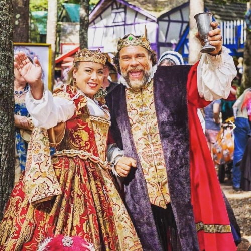 A man in royal attire and a woman in a colorful gown wave cheerfully at a festival, surrounded by medieval-themed decorations.