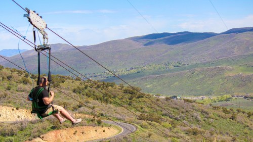 A person zip-lining over a scenic landscape with rolling green hills and a winding road below.