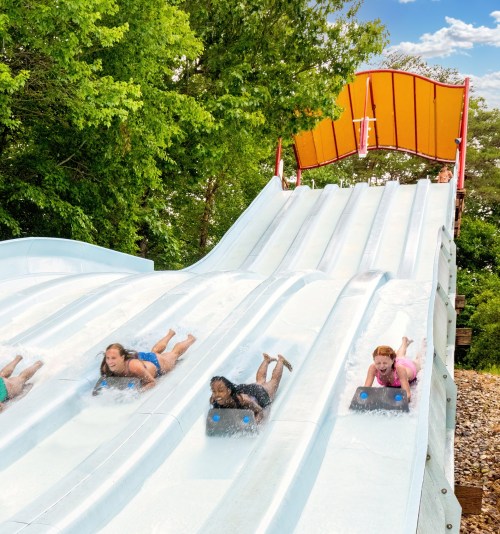 Four children slide down a water slide, splashing into a pool below, surrounded by trees and a bright blue sky.