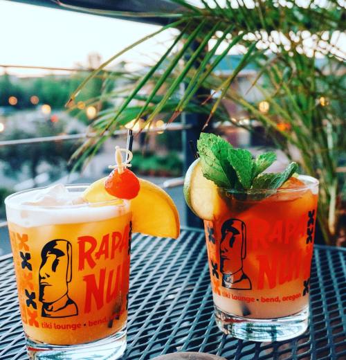 Two colorful cocktails in glasses with "Rapa Nui" branding, garnished with fruit and mint, on a patio table.