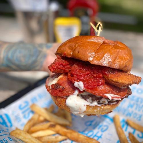 A close-up of a stacked burger with meat, sauce, and toppings, served with a side of fries on a wooden table.