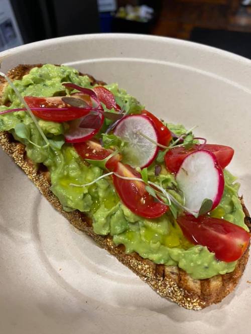 A slice of toasted bread topped with mashed avocado, cherry tomatoes, radish slices, and microgreens, drizzled with olive oil.