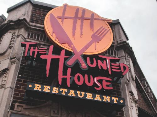 Sign for "The Haunted House Restaurant" featuring a knife and fork design, set against a cloudy sky.