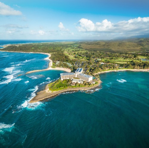 Aerial view of a coastal resort surrounded by turquoise waters and lush green landscapes under a blue sky.