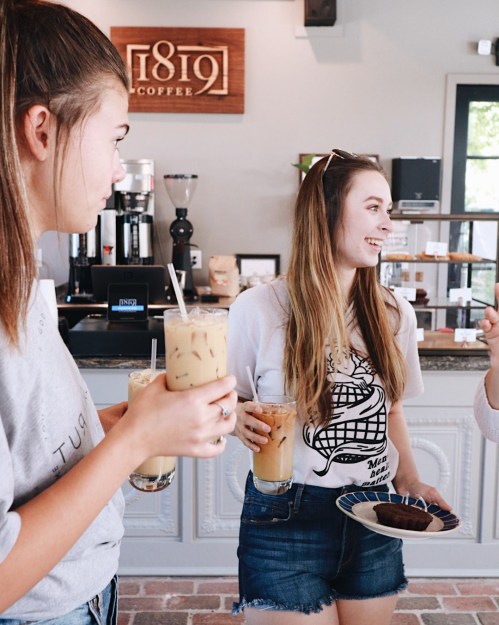 Two young women in a café, one holding iced coffee and a plate of brownies, chatting and smiling.