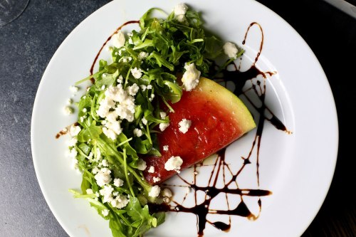 A plate featuring a slice of watermelon topped with arugula and crumbled feta, drizzled with balsamic glaze.