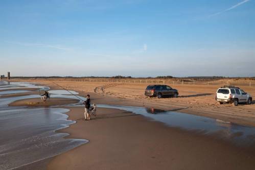 Two people fishing on a sandy beach with vehicles parked nearby and a calm sea under a clear blue sky.