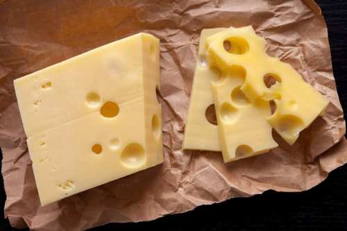 A block of Swiss cheese with holes, partially cut into slices, resting on crumpled brown paper.