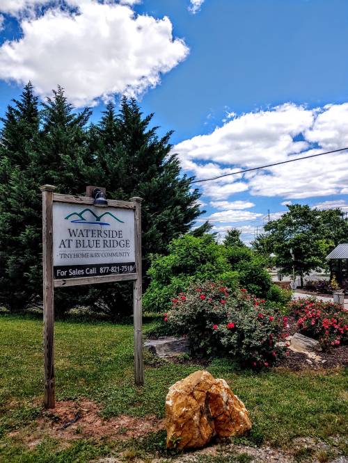 Sign for "Waterside at Blue Ridge" surrounded by greenery and a rock, under a blue sky with clouds.