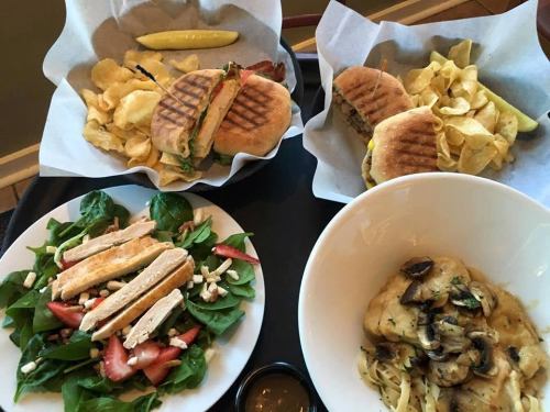 A table with two sandwiches, chips, a salad with chicken, and a bowl of pasta with mushrooms.