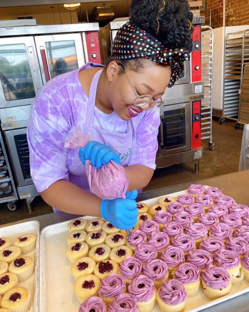 A woman in a purple shirt and gloves decorates cupcakes with purple frosting in a bakery.