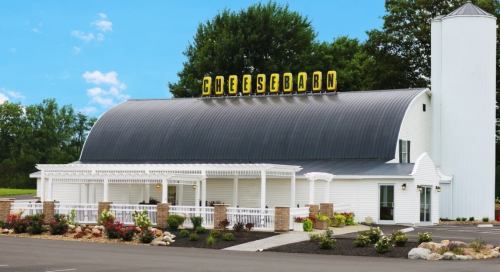 A white barn-style building with a curved roof, featuring a sign that reads "Cheese Barn" and surrounded by greenery.