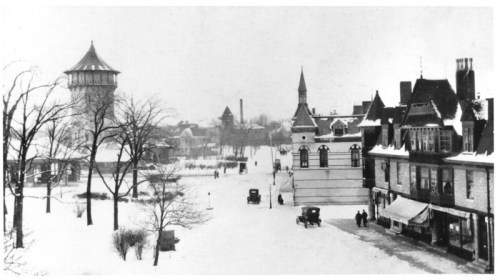 Black and white winter scene of a snowy town square with historic buildings and vintage cars.