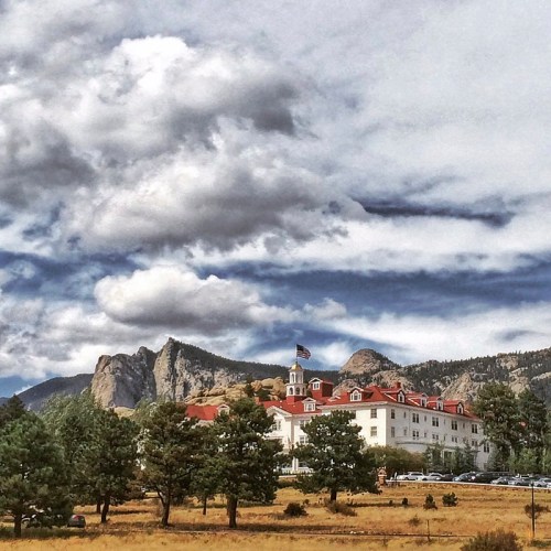 Historic hotel with red roofs, surrounded by trees and mountains under a cloudy sky.