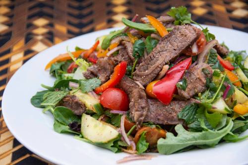A vibrant salad topped with grilled beef, mixed greens, cherry tomatoes, and colorful bell peppers on a white plate.