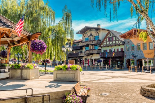Charming town square with colorful flowers, historic buildings, and a clear blue sky. American flag visible.