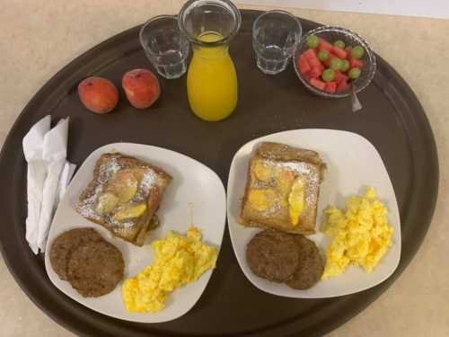 A tray with two plates of French toast, scrambled eggs, sausage, fruit, and a glass of orange juice.