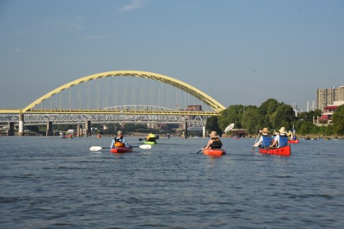 A group of kayakers paddles on a river with a yellow arch bridge in the background under a clear blue sky.