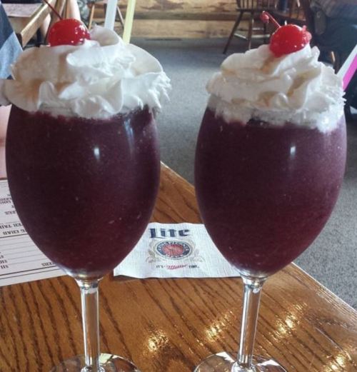 Two tall glasses filled with a purple slushy drink, topped with whipped cream and a cherry, on a wooden table.