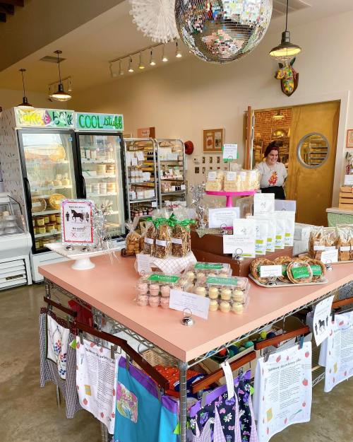 A cozy shop interior with a pink table displaying treats, a fridge, and a smiling employee at the entrance.